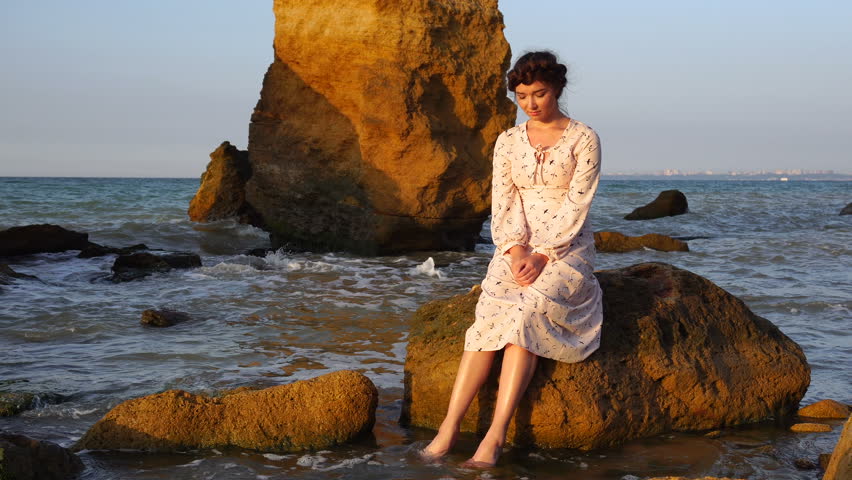 Adorable Caucasian Brunette girl in Light Pink Dress Sit on the Rock in the Sea at Sunrise Time