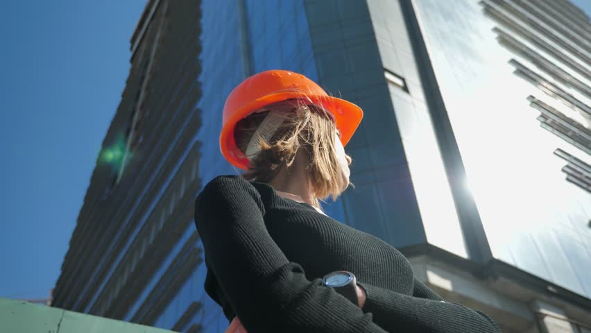 Young girl builder with hemlet is standing with arms crossed in daytime in summer, building concept, urban concept, building on background, bottom view