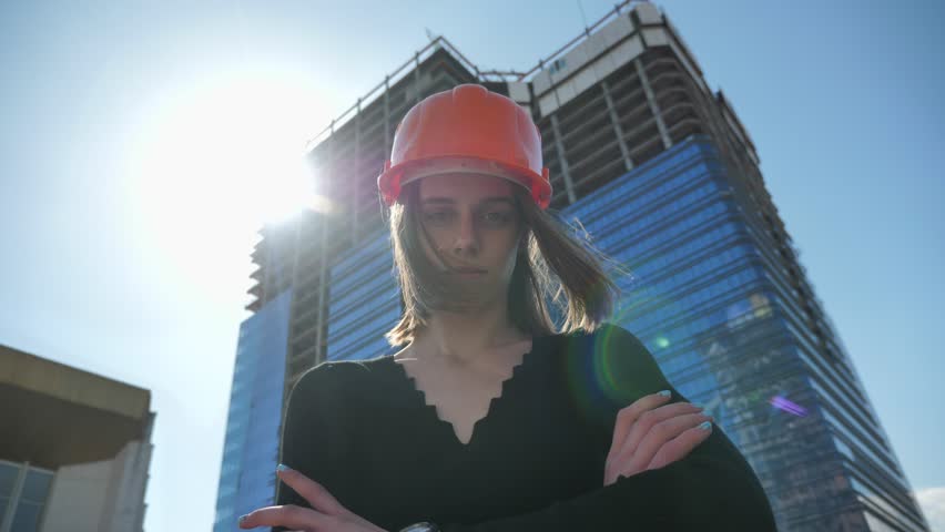 Young strong girl builder with hemlet is standing with arms crossed in daytime in summer, building concept, urban concept, building on background, bottom view