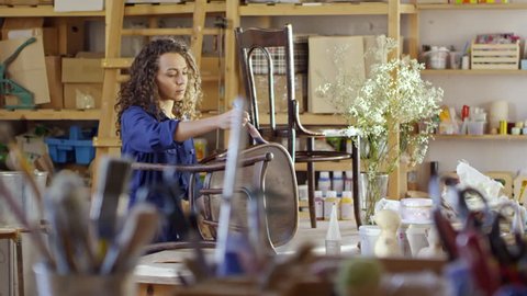 Pretty young woman with curly hair using brush to apply varnish on old wooden chairs in carpentry workshop