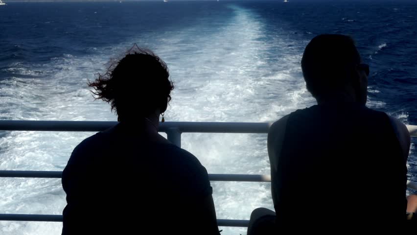 Rear shot from a silhouette couple sitting together at the end of a ship looking at the sea and V shaped pattern of Kelvin wake