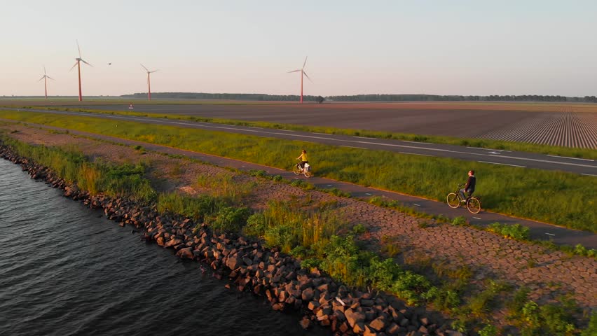Couple enjoying a bicycle ride on a bike path along shore of Dutch polder Flevoland, with windmill park and fields on the background, at sunset. Royalty-Free Stock Footage #1013503172