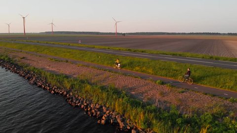 Couple enjoying a bicycle ride on a bike path along shore of Dutch polder Flevoland, with windmill park and fields on the background, at sunset.