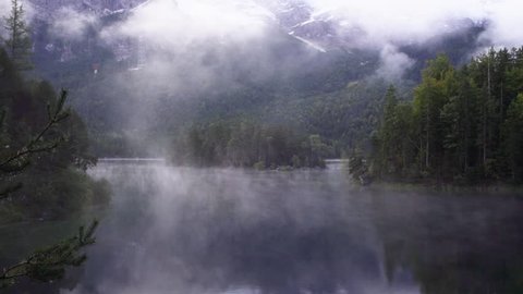Fog drifts over an alpine lake surrounded by forest trees and mountains early in the morning