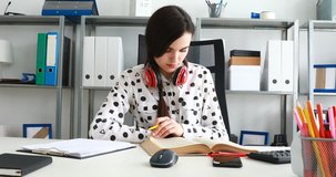 woman with red headphones on shoulders thumbing book and writing in pencil on paper.