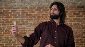 Young attractive brunette man with beard is making video on smartphone, fooling around, laughing, brick background, communication concept