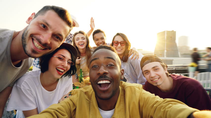 Point of view shot of young people multiethnic group taking selfie and holding camera, men and women are looking at camera, smiling and posing with drinks at rooftop party. Royalty-Free Stock Footage #1013525135