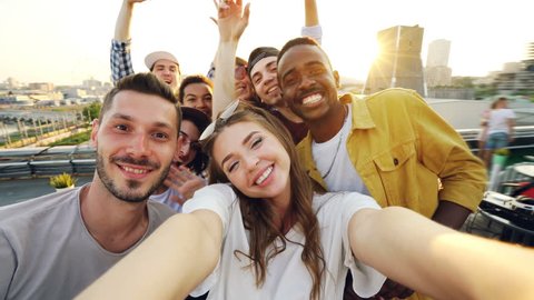 Attractive young woman is taking selfie with friends on rooftop, girl is holding camera and posing while her mates are having fun making funny faces and gestures.