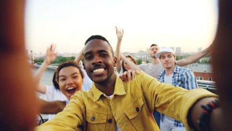 Point of view shot of handsome African American man partying with friends on rooftop, dancing and having fun with drinks. Sky and beautiful city is visible.