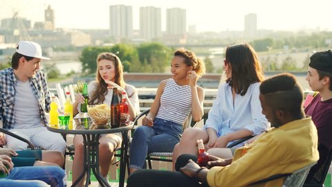 Pretty young girl is telling story to her friends multiethnic group sitting at table on rooftop with food and drinks, beautiful view of city is in background.