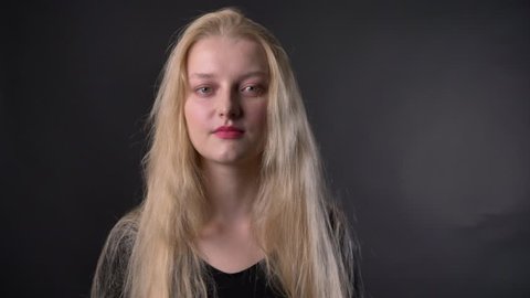 Young beautiful tired blond girl with pink lipstick is watching at camera, grey background