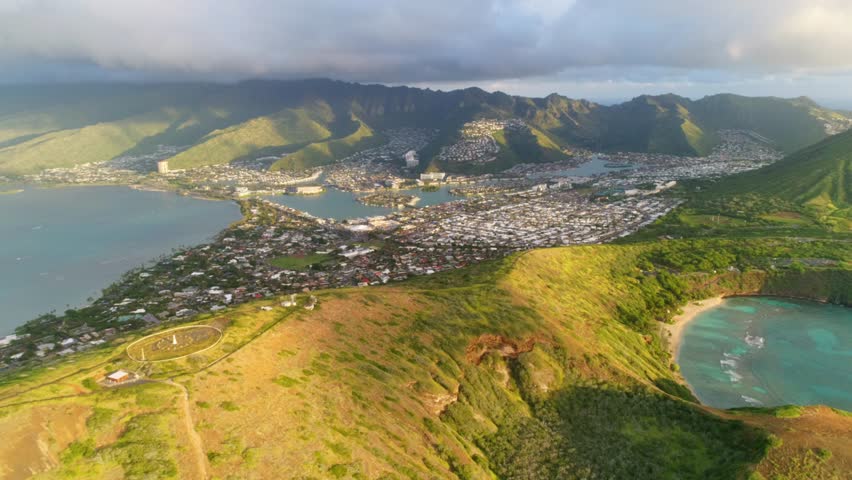 Aerial shot of an urban town nestled into a valley on a tropical island. Mountains, hills, craters, clouds and a bay populate the landscape. Shot during sunrise. 