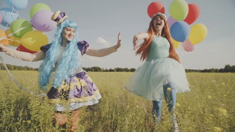 Two cheerful girls in bright costumes of princesses and with balloons are having fun in the field. HD
