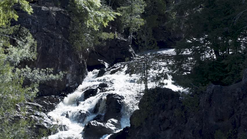 Heavy pouring waterfall panning downward to the drop off into a river. Setting is nestled in the woods showing a rocky cliff and scenic forest located in Northern Wisconsin.