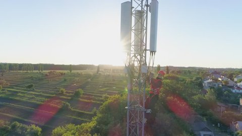 aerial view of telecommunications tower, technician in a safety vest and hard hat uses mobile phone on top of cellular antenna