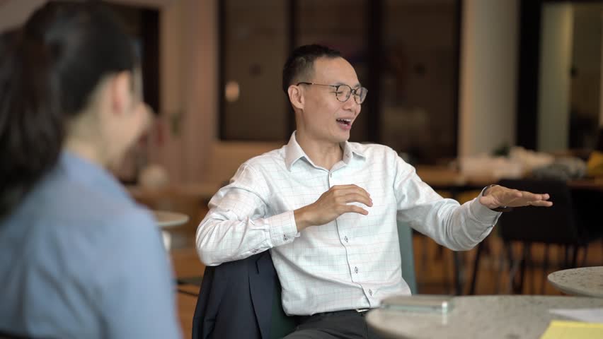 A Chinese Asian business man is sitting and explaining a concept animatedly as he smiles to his team in the office. He is professionally dressed in shirt and pants and looks competent and mature.  Royalty-Free Stock Footage #1013585711