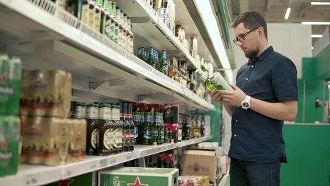 Russia Togliatti - June 2018: Male visitor of a supermarket is a holding in hands glass bottle and metal can with beer. He is reading inscription on labels in a store