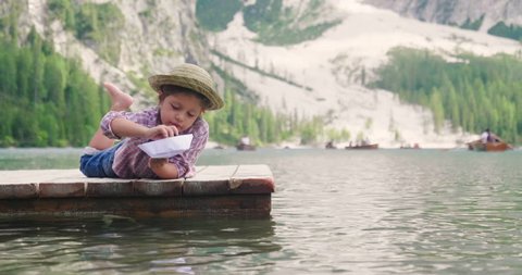 A little girl on the pier by the lake plays with a paper boat making her surf and blowing to make her travel. 