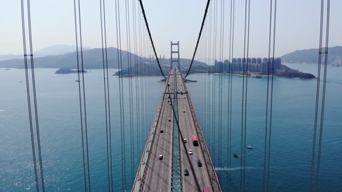 Impressive lengthwise aerial perspective of large suspension bridge, camera fly along road, between cables. Light traffic on highway, modern Lantau Link at Hong Kong city. Bridge span across channel