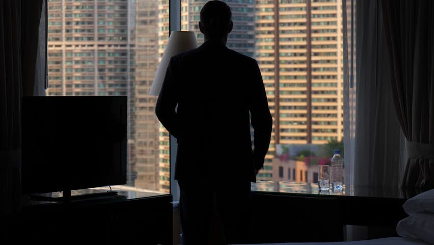 Business man stay with hands akimbo, silhouette against room window. Man wear black jacket, put arms on hips and look out to city, Then turn around and walk away