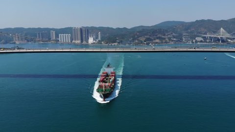 Big container ship pass Ma Wan channel, move towards from under Tsing Ma bridge, aerial shot. Large cargo carrier perform ride at Hong Kong shipway, auto traffic seen on highway deck of bridge