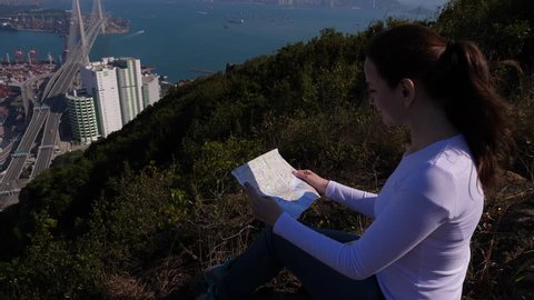 Woman look to Hong Kong city paper map, sitting on rock at high point. Tourist woman discover Tsing Yi island and scenic viewpoints of famous Asian city, rest after walking severe trekking path