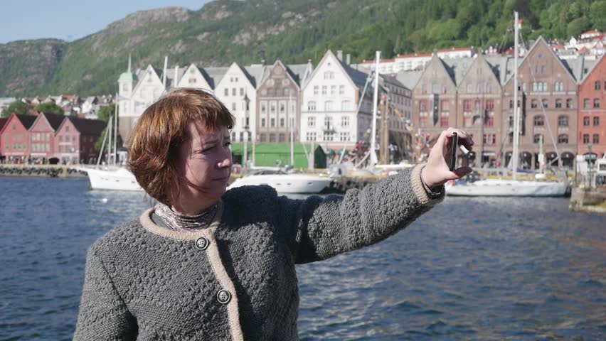 Middle aged woman makes selfie looking at the smart phone on Bergen old town background