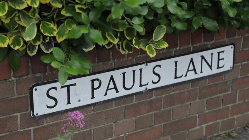 A vintage road sign on a brick wall with flowers and shrubbery growing around it.