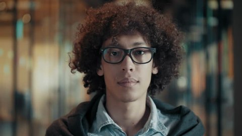 Close up face of tranquil calm african young male in glasses black guy with dark curly hair portrait of creative young man in stylish clothes in modern start up office quietly watching and blinking