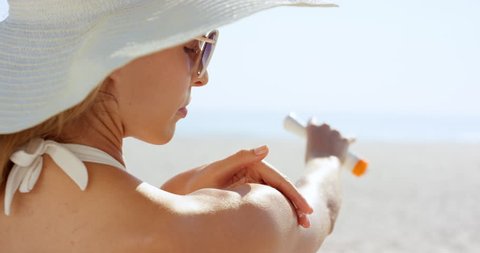 Close up of woman applying sunscreen to shoulder on tropical beach wearing one piece white bathing suite and white hat RED DRAGON