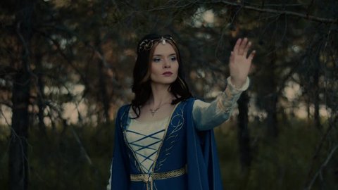 Medieval witch is conjuring alone in a forest in summer. She is moving hand from up to down and crossing hands on a chest