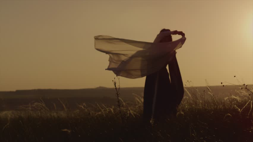 Medieval girl is whirling a cloth around her head. She is standing against sunset sun in a lawn, view on her silhouette