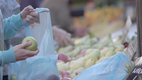 Slow Motion Shot Of Male And Female Customers Hands Choosing Green Apples And Bagging Them Into Blue Plastic Bags At A Street Fruit Market Stall, Supplying Plastic Carrying Bags To Customers.