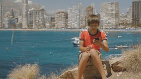 15-years-old teen boy makes smartphone video call on skyscrapers skyline background. Ship passing in the sea bay behind