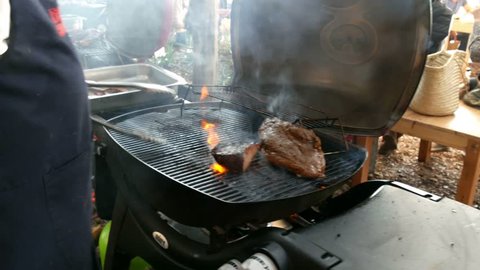 Barbecue grill with lid open, meat on grid being cooked, man holding tongs holds picks up, taps & turns meat with fat dripping into fire causing a flare of orange flames