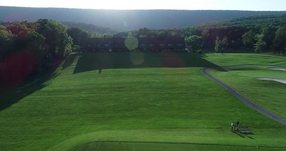 Aerial camera ascending from golf course to show the lodge on a green golf course set against a setting sun.
