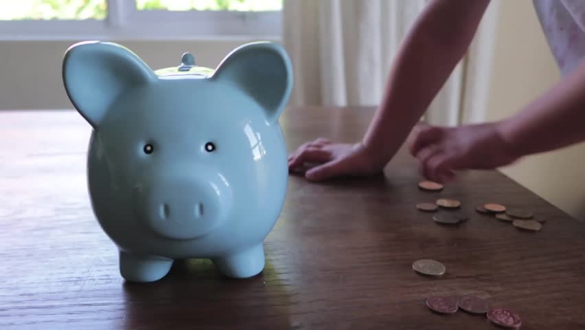 close up footage of a youg childs hand putting money into a blue piggy bank