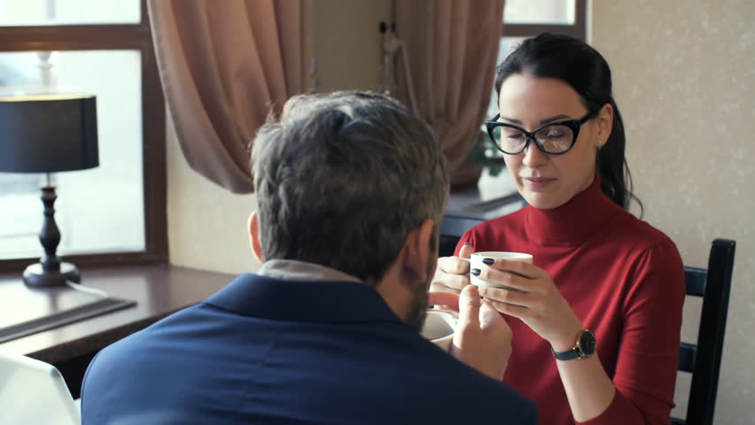 Medium shot of attractive young woman in glasses listening to male friend or colleague and laughing when enjoying coffee during break from work
