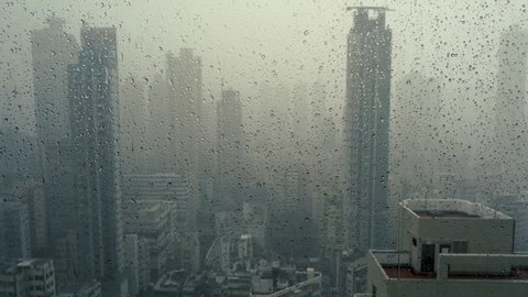 Rain falling on window in skyscraper apartment. Dark and stormy rainy day in big city life. View of dreary Hong Kong cityscape whilst rain drops fall on the window. Abstract somber moody background.