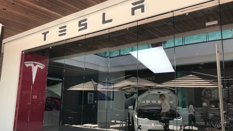 LOS ANGELES, JULY 7TH 2018: Steady shot of the Tesla car dealership at the Westfield shopping mall in Century City, with people walking past and a woman closing a car's hood, seen through glass panels