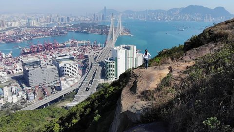 Woman stay on rock, admire urban panorama from view point, aerial shot. Large bridge and container terminals seen ahead, industrial shore of Tsing Yi island from above. Camera fly over hiker
