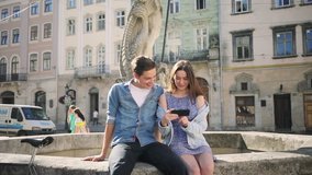 Young cute Caucasian couple in love sitting beside each other on background of beautiful fountain. Girl and guy looking funny video on social media. Outdoors.
