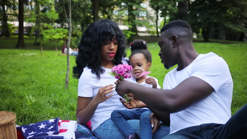 Nice African family on picnic in wonderful greeen park. Father giving bouquet of pink flowers to little pretty girl. Young people sitting on blanket. Outdoors. | Shutterstock HD Video #1013716394