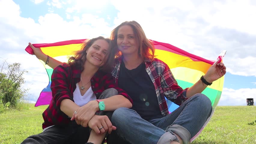 Two young women are sitting on a background of the rainbow flag. The sun is shining brightly, LGBT rights, lesbian family.