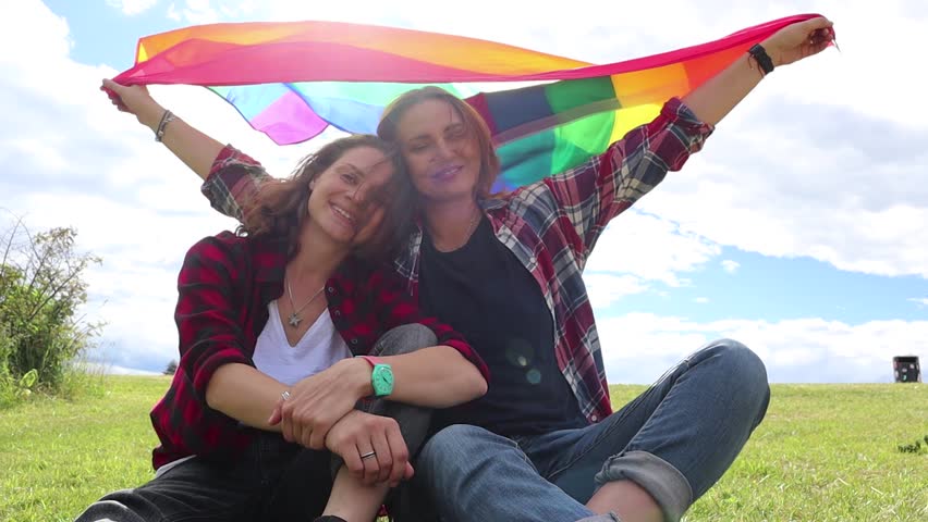 Two young women are sitting on a background of the rainbow flag. The sun is shining brightly, LGBT rights, lesbian family. Royalty-Free Stock Footage #1013725067