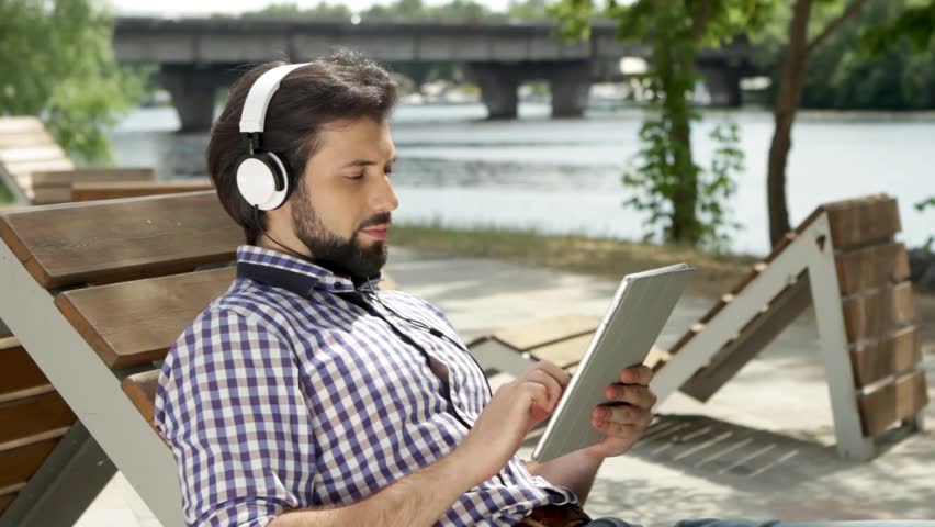 Guy is lying on bench and listening to music through headphones. Also he is holding tablet and typing on screen. Man is looking at screen and moving his fungers. He slides down and up. Guy is smiling.