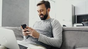 Smiling bearded man using smartphone while sitting on couch with laptop computer at home