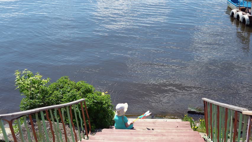 Little girl in a blue dress and hat is sitting on a wooden pier enjoying sea view on a sunny day. The child holds a colorful pinwheel in his hands,back view.