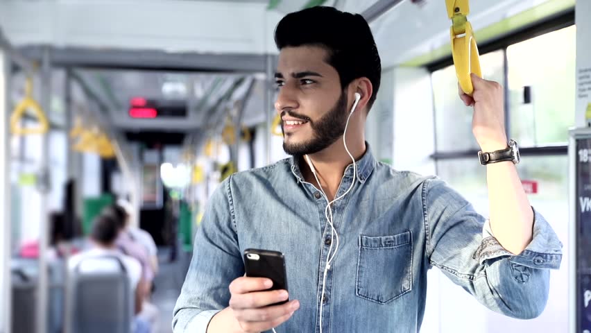 Close up view of Handsome Man Standing on the Public Transport. Interior of Crowded Tram. Man Types on his Mobile Phone. Listening to Music with his Headphones. Luxurious Wristwatch. Casual Clothing.
