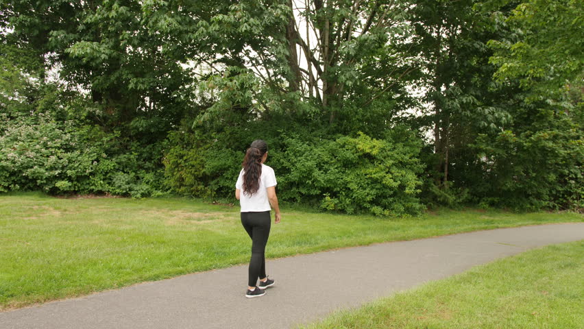 A Dark Haired Latina Woman Stretching in Yoga Pants and a White Shirt Before Starting Her Run in a Lush Green Park